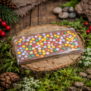 Czekolada mleczna z kolorowymi drażetkami zapakowana w folię, ułożona na drewnianym krążku w leśnej aranżacji