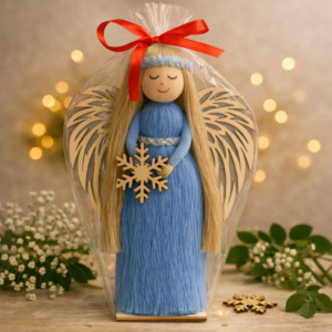 Ręcznie wykonana figurka anioła w niebieskiej sukni z drewnianymi skrzydłami i płatkiem śniegu na tle świątecznych światełek.